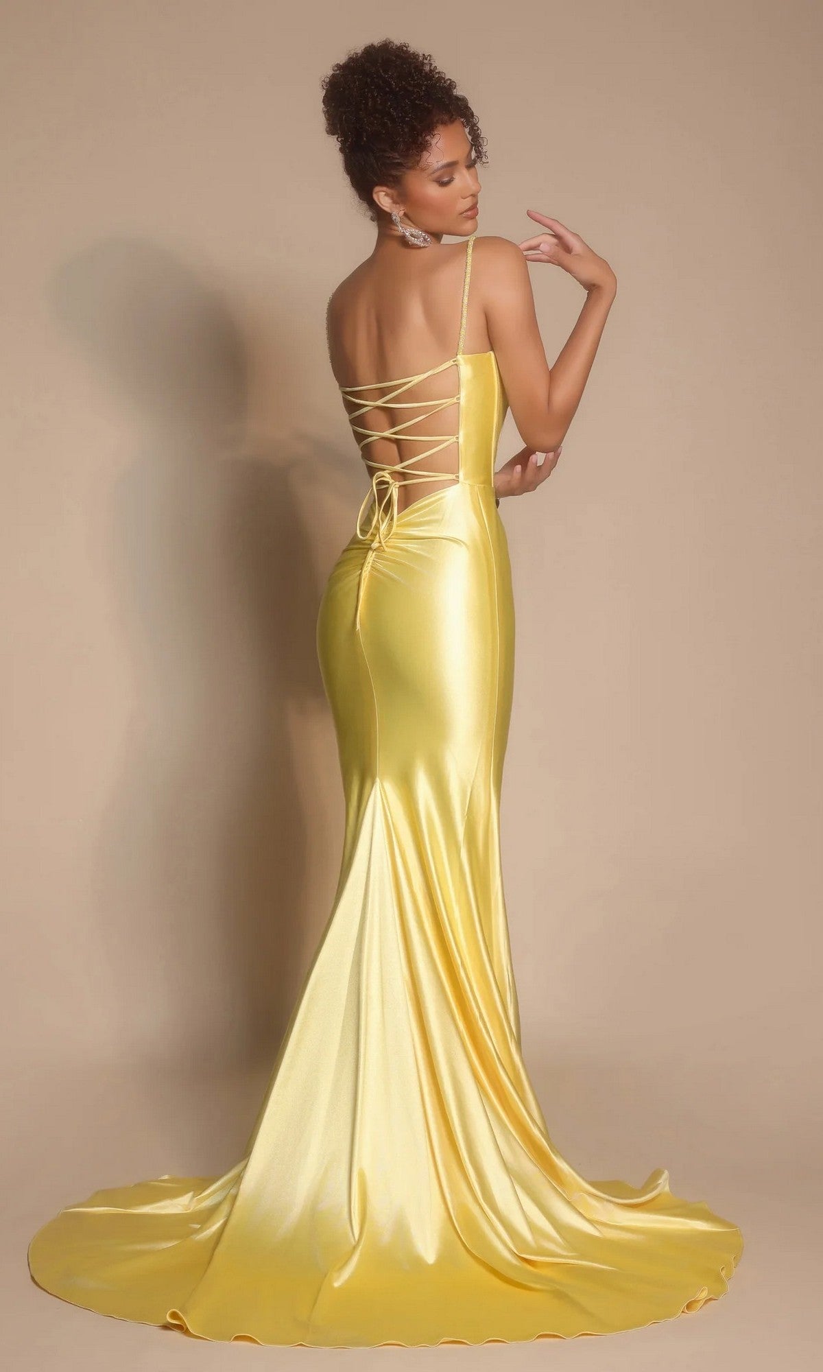 Woman wears the Portia and Scarlett XO042 long formal dress, a shiny yellow satin gown with a corset bodice and lace-up back, posing before a plain beige background.